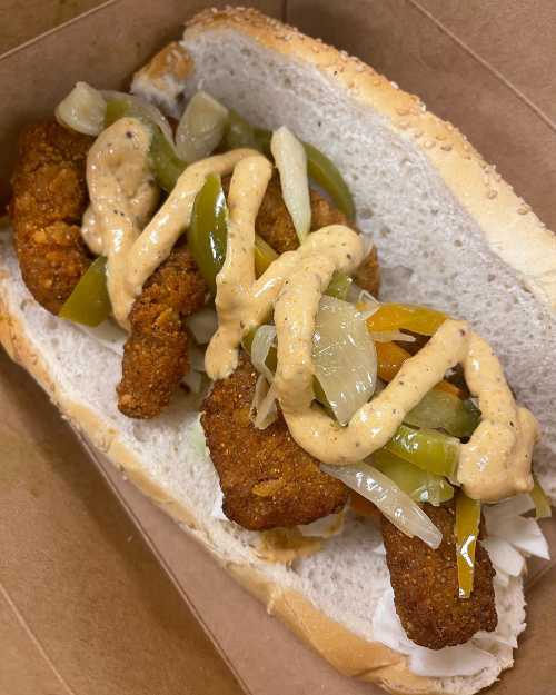 A sandwich with crispy fried pieces, sautéed onions, and green peppers, drizzled with a creamy sauce on a sesame seed bun.