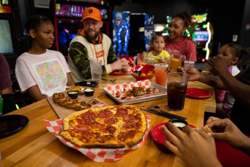 A group of people enjoying pizza and drinks at a lively restaurant with arcade games in the background.