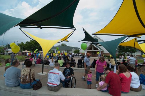 A community event with colorful canopies, people sitting, and balloons, creating a festive atmosphere.