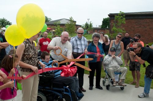 A group of people gathers for a ribbon-cutting ceremony, with balloons and a large pair of scissors in the foreground.