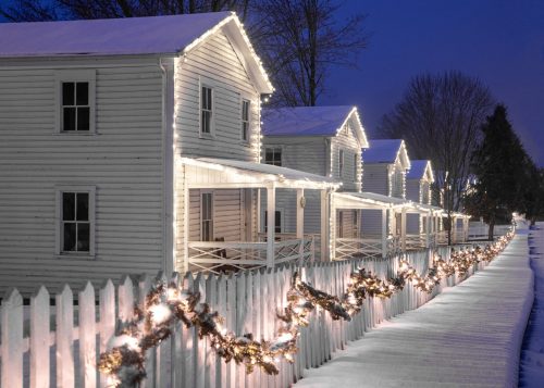 Snow-covered houses adorned with lights and garlands, lined along a white picket fence in a serene winter evening.