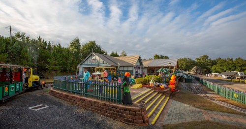 A colorful outdoor scene featuring a train, decorations, and a building surrounded by trees under a cloudy sky.