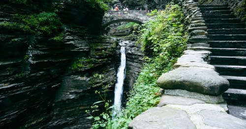 A waterfall cascades down rocky cliffs, with a stone bridge above and lush greenery surrounding the scene.