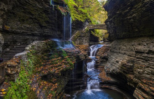 A serene gorge with cascading waterfalls, surrounded by rocky cliffs and autumn foliage, featuring a stone bridge above.