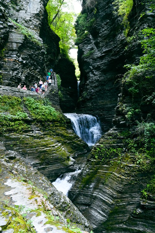 A narrow canyon with lush greenery and a waterfall, with people walking along the rocky path beside the water.