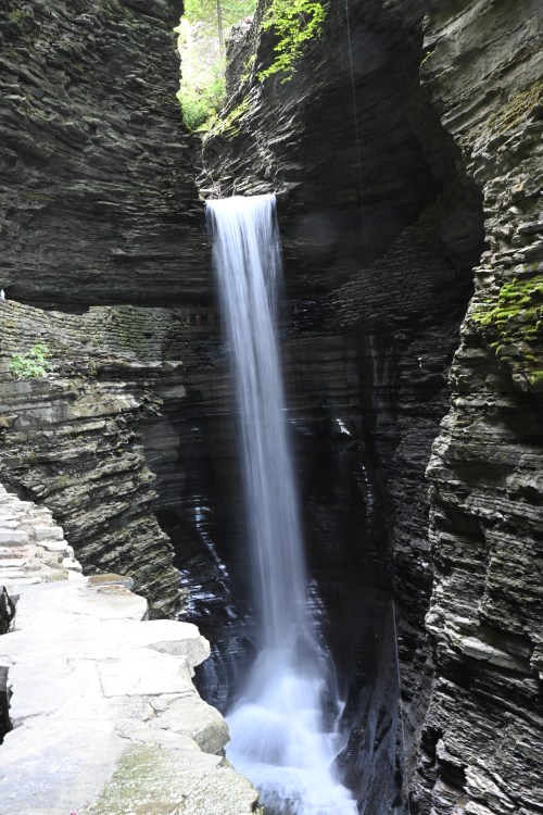 A serene waterfall cascades down rocky cliffs, surrounded by lush greenery and a stone pathway.
