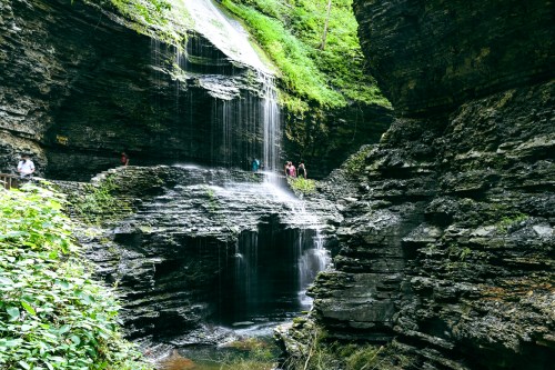 A serene waterfall cascades over rocky cliffs, surrounded by lush greenery and visitors exploring the area.