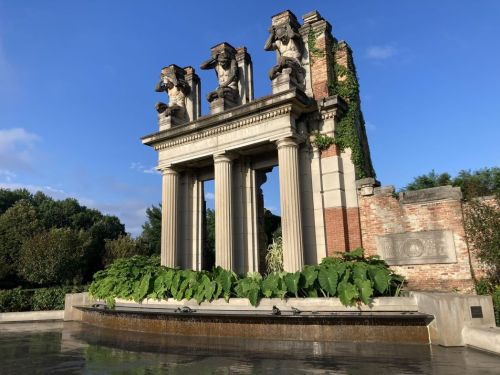 A historic stone structure with four lion sculptures, surrounded by greenery and a reflecting pool under a blue sky.