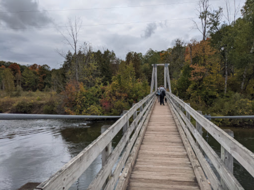 A wooden bridge spans a river, surrounded by trees with autumn foliage under a cloudy sky. Two people walk across it.