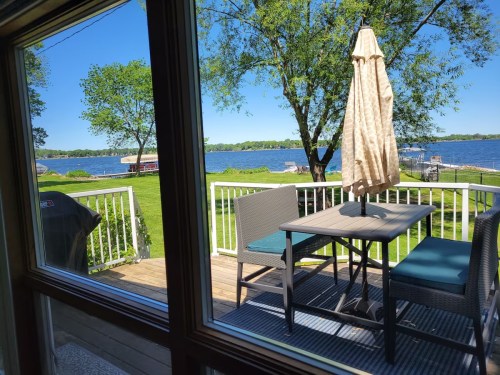 View from a window showing a patio with a table and chairs, overlooking a lake and green landscape on a sunny day.