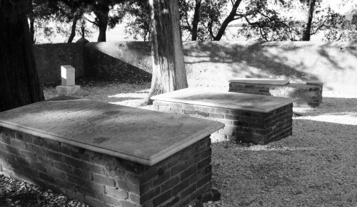 Black and white image of stone graves surrounded by trees and gravel, with a serene, shaded atmosphere.