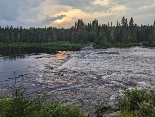 A serene river flows through a forested landscape, with a sunset peeking through the trees and clouds above.