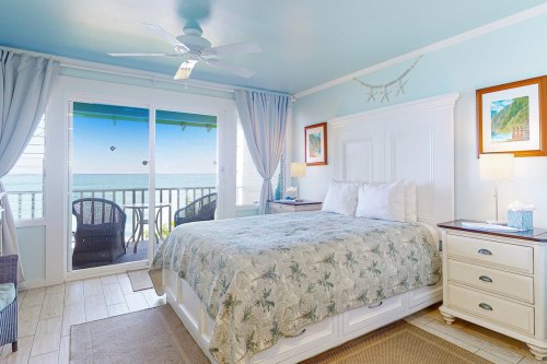 Bright coastal bedroom with a queen bed, light blue walls, and a view of the ocean through sliding glass doors.