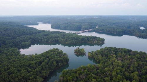 Aerial view of lush green forests surrounding calm lakes and a bridge in the distance under a cloudy sky.