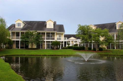 A serene pond with a fountain surrounded by green grass and two-story buildings with balconies in a peaceful setting.