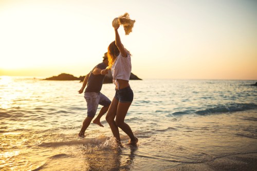 A couple joyfully dances in the shallow water at sunset, with waves gently lapping around them.