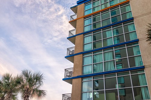 A modern building with large windows and balconies, surrounded by palm trees under a cloudy sky.