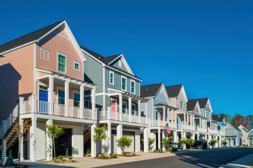 Colorful, modern houses with porches line a sunny street, featuring greenery and clear blue skies.