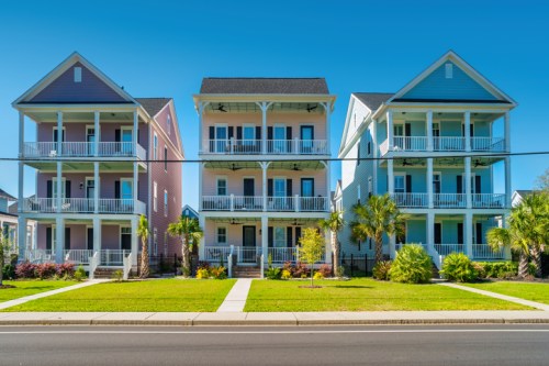 Colorful multi-story houses with balconies, lined up along a sunny street, surrounded by greenery and palm trees.