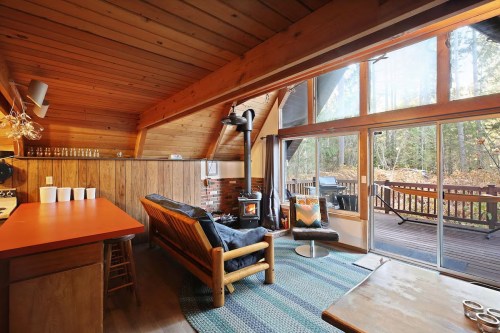 Cozy cabin interior with wooden beams, a seating area, and large windows overlooking a deck and forest.