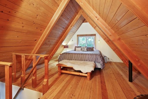 Cozy attic bedroom with wooden walls, a large bed, and a window overlooking greenery. Warm, rustic decor.