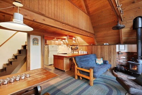 Cozy wooden cabin interior with a living area, kitchen, and staircase, featuring a sofa and a wood stove.