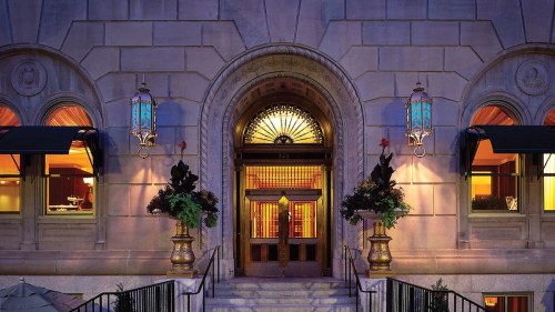 Elegant entrance of a building at night, featuring ornate lanterns and decorative plants. Warm lights glow from inside.