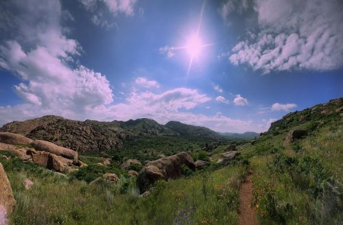 A panoramic view of a lush green landscape with rocky hills under a bright blue sky and fluffy white clouds.