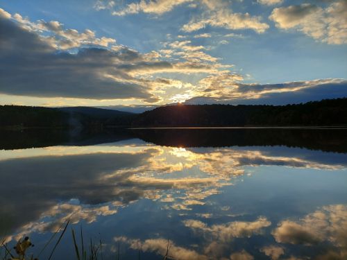 A serene lake at sunset, reflecting colorful clouds and mountains in calm water, surrounded by lush greenery.