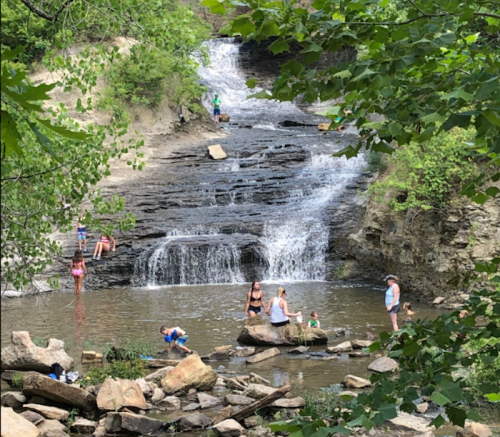A scenic waterfall surrounded by greenery, with people enjoying the water and rocks in a natural setting.