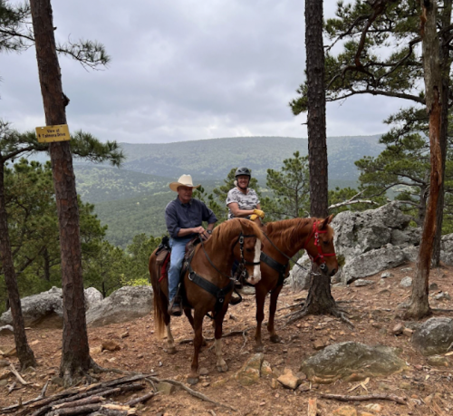 Two people on horseback in a forested area, with mountains in the background and a cloudy sky above.