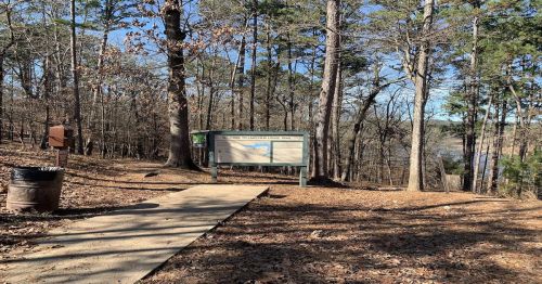 A trailhead sign surrounded by trees, with a paved path leading through a wooded area.