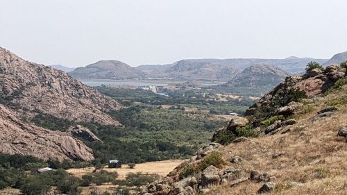 A scenic view of rolling hills and a distant dam, surrounded by greenery and rocky terrain under a hazy sky.
