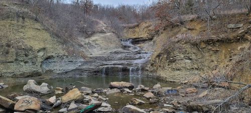 A rocky landscape featuring a small waterfall cascading down a cliff into a pool, surrounded by bare trees and shrubs.