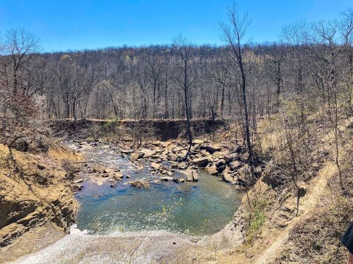 A serene river flows through a rocky landscape, surrounded by bare trees under a clear blue sky.