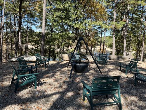 A serene outdoor space with green chairs arranged around a fire pit, surrounded by trees and a peaceful landscape.
