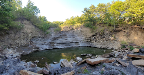 A serene rocky landscape with a small pond surrounded by steep, layered cliffs and lush green trees.