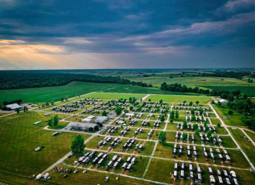 Aerial view of a large RV park with numerous parked vehicles, surrounded by green fields and a cloudy sky.