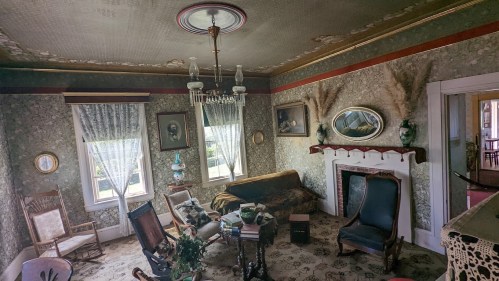 A vintage living room with floral wallpaper, antique furniture, and large windows, featuring a chandelier and decorative mirrors.