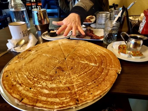 A large, round dosa on a plate with condiments and a hand reaching out, set on a table with various breakfast items.