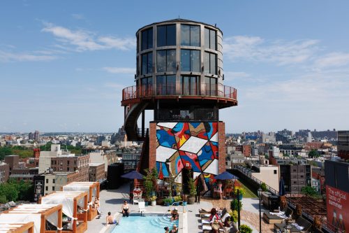 A rooftop pool area with a colorful mural and a unique tower structure, overlooking a city skyline on a sunny day.