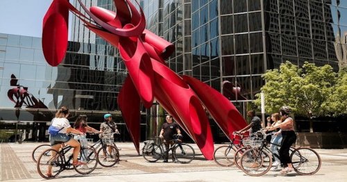 A group of cyclists gather near a large red sculpture in an urban setting with glass buildings in the background.