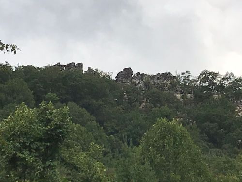 Ruins of a stone structure atop a green, wooded hill under a cloudy sky.