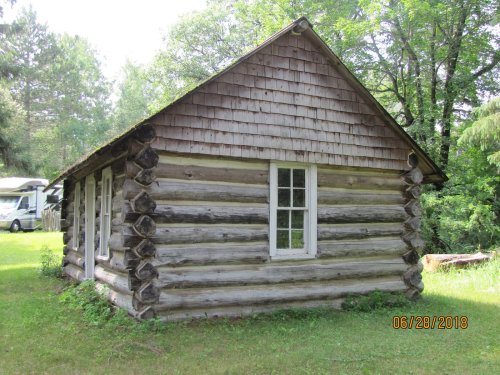 A rustic log cabin with a gabled roof, surrounded by greenery and trees, featuring a window on the side.