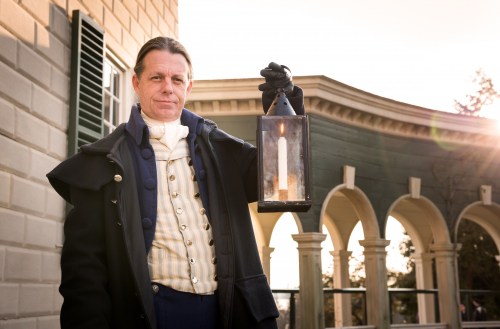 A man in historical attire holds a lantern, standing outside a building with arches in the background.