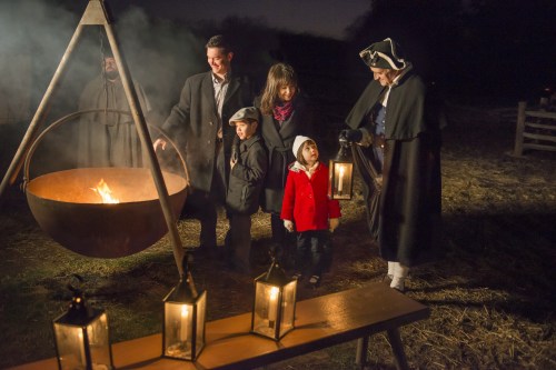 A family gathers around a large cauldron over a fire at night, with lanterns illuminating the scene.