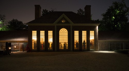 A historic building illuminated at night, with people visible through large windows and trees in the background.