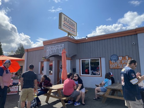A busy restaurant exterior with people sitting outside, enjoying the sunny day and a sign that reads "Breakfast."