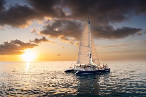 A sailboat with people on board glides across calm waters at sunset, with vibrant clouds in the sky.