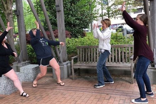 Four people playfully posing under a wooden structure in a park, with trees and a bench in the background.